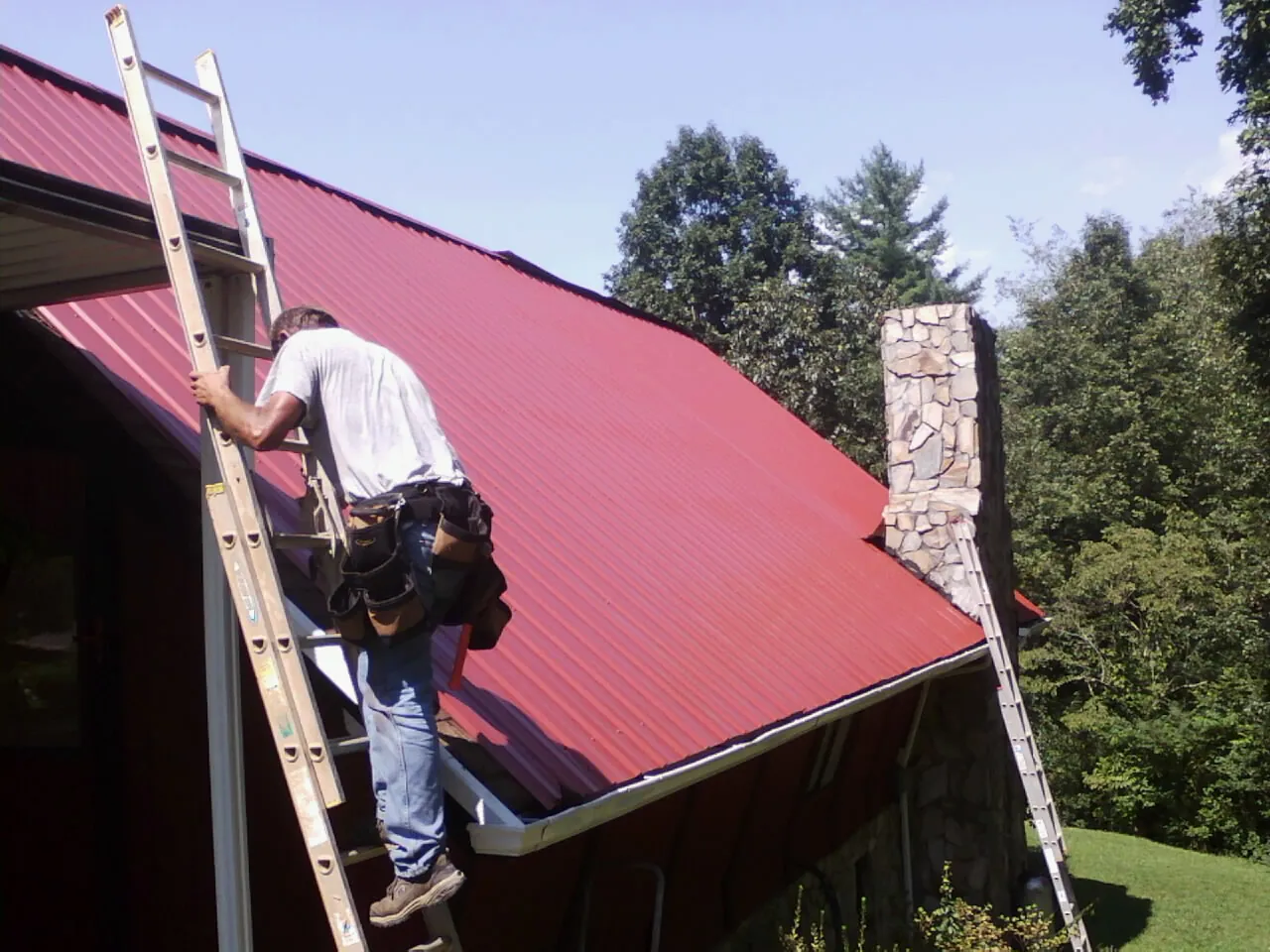 Roofer on a ladder beside a red metal roof and stone chimney — Soffit & Fascia Repair by Roofing Brothers in Los Lunas